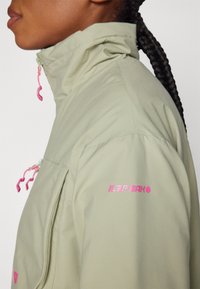 Veste vert clair avec un col montant, poches zippées et cordons roses. Présente un logo rose sur la manche. Texture lisse.
