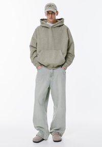 Hanorac supradimensionat de culoare verde deschis, cu un buzunar în față, combinat cu blugi de denim deschiși, cu picior larg, și pantofi slip-on gri. Săcărici are un text imprimat.