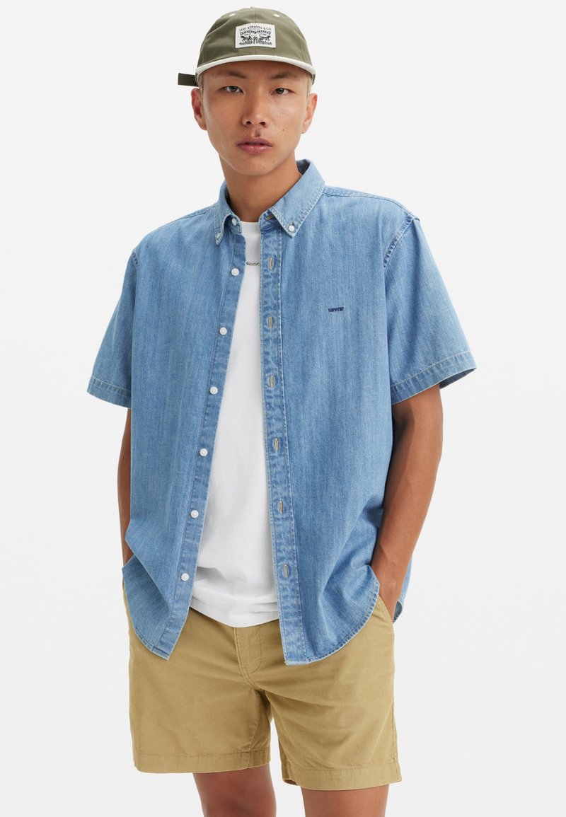 Light blue denim short-sleeve shirt with a button-down collar, worn over a white T-shirt, paired with khaki shorts.