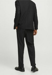 Czarny garnitur, składający się z jedno-zapięciowej marynarki oraz dopasowanych spodni, o gładkim wykończeniu i prostym kroju nogawki. Widoczne czarne buty.