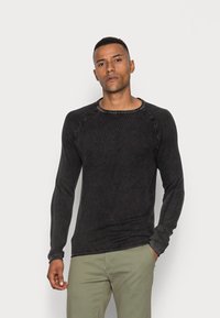 Schwarzes Langarm-T-Shirt aus strukturiertem Stoff mit Rundhalsausschnitt und kontrastierenden Nähten, kombiniert mit hellgrünen Hosen.