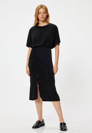 Zwarte jurk met korte mouwen, gathered textuur bij de taille, een diepe split aan de voorkant en een gladde stof. Gecombineerd met zwarte schoenen.