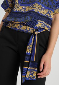 Ein blauer und gold gemusterter Oberteil mit gebundenem Bund, das aufwendige Designs und schwarze Kontrastelemente aufweist, kombiniert mit schwarzen Hosen.