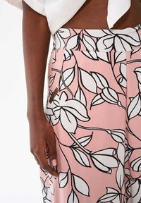 Spodnie w kwiatowy wzór z różowym tłem i czarno-białym wzorem liści. Mała brązowa metka z logo jest wszyta w pasek.