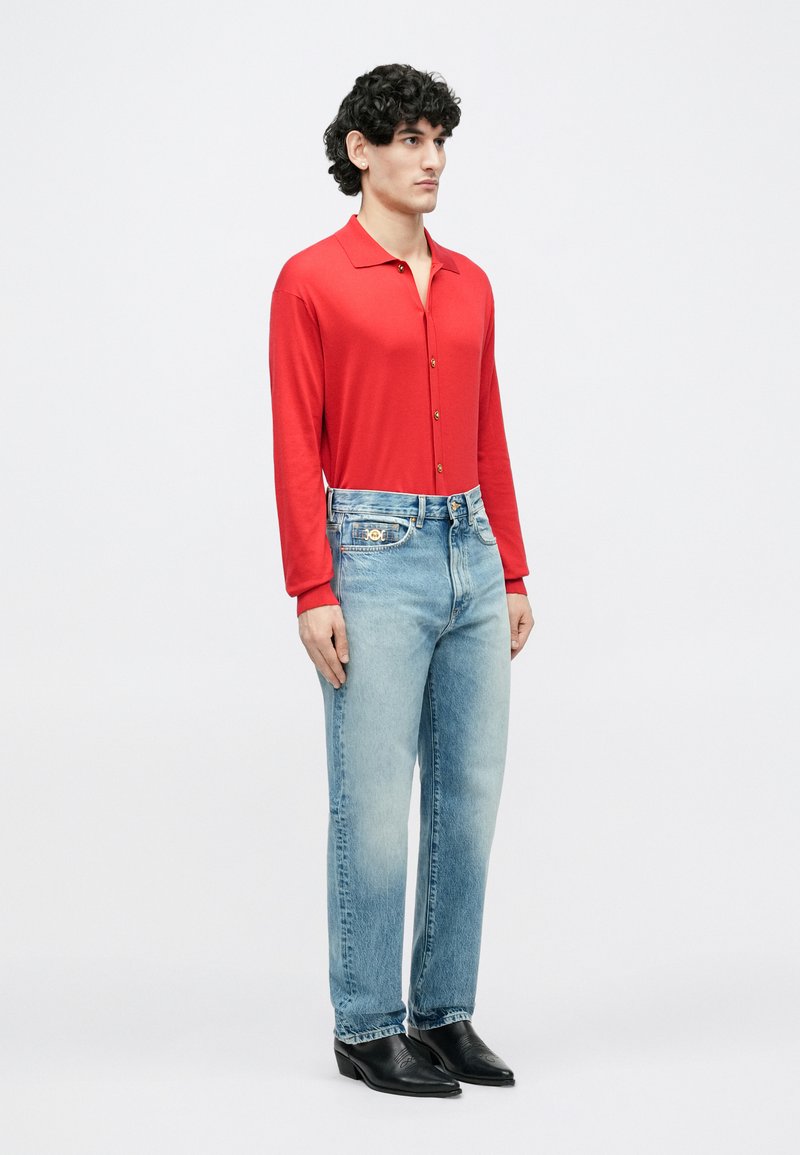 Man met krullend haar, gekleed in een rood shirt met lange mouwen, lichtblauwe spijkerbroek en zwarte leren schoenen met een puntige neus, staand tegen een effen achtergrond.