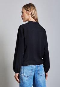 Blusa nera in tessuto con maniche lunghe, orlo arrotondato e leggere pieghe ai polsini, abbinata a jeans in denim azzurro chiaro.