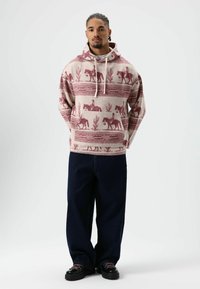 Sweat à capuche beige clair avec des motifs rouges de style western représentant des chevaux et des cactus, poche avant kangourou, capuche avec cordon de serrage, accompagné d'un jean baggy bleu foncé.