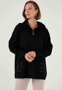 Pull noir avec un col montant et une demi-fermeture éclair, texture côtelée et accents de clous décoratifs sur deux poches avant.
