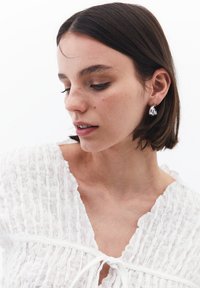 Blusa bianca con texture, scollo a V e dettaglio con fiocco, indossata con orecchini a goccia argentati. Il tono della pelle uniforme esalta il design minimalista.