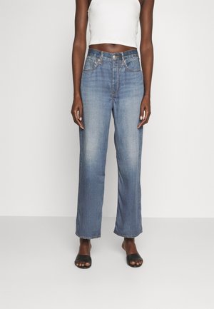 Person med lyseblå jeans med lige ben, hvid cropped tanktop og sorte åbne slid-sandaler mod hvid baggrund.