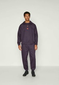 Marineblauwe tracksuit van gladde stof; voorzien van een hoge kraag, geborduurd logo en elastische mouwen; gedragen met zwarte sneakers.