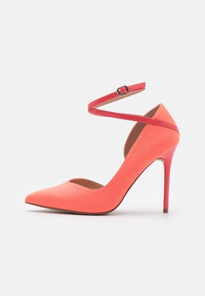 Zapato de tacón alto de ante coral con punta afilada, correa al tobillo con hebilla, textura suave y tacón de aguja delgado.