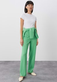 Witte kortarmige fitted top gecombineerd met hoogsluitende groene broeken met een knoopdetail in de taille. Beige puntige flats.