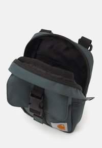 Bolsa crossbody de lona verde-escuro com aba, alça ajustável e fecho de fivela. O interior apresenta forro de tecido preto e bolsos.