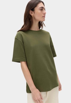 Kvinde iført en olivengrøn T-shirt med korte ærmer og cremefarvede bukser, vendt mod venstre foran en ensartet lysegrå baggrund.