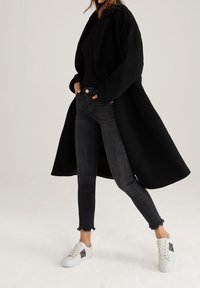 Manteau noir à larges revers porté sur un pull noir, associé à un jean foncé avec des ourlets effilochés et des baskets blanches avec des accents noirs.