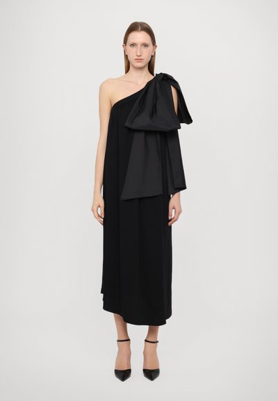 Robe noire sans manches avec une épaule, ornée d'un grand nœud en tissu. Silhouette fluide avec une texture lisse, longueur mi-genou.