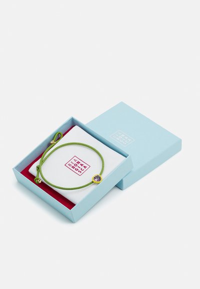 Zeleno pletena zapestnica s purpurnim dragim kamnom, predstavljena v svetlo modri škatli z rdečim notranjostjo in belo blazinico, na kateri je prikazana blagovna znamka.