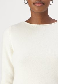 Pull en tricot blanc côtelé avec un col rond et des manches longues, présentant une texture subtile et des détails de design minimaux.