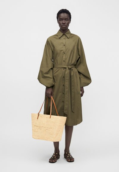 Femme en robe boutonnée vert olive avec taille nouée et manches bouffantes, tenant un sac fourre-tout beige tissé et portant des sandales foncées.