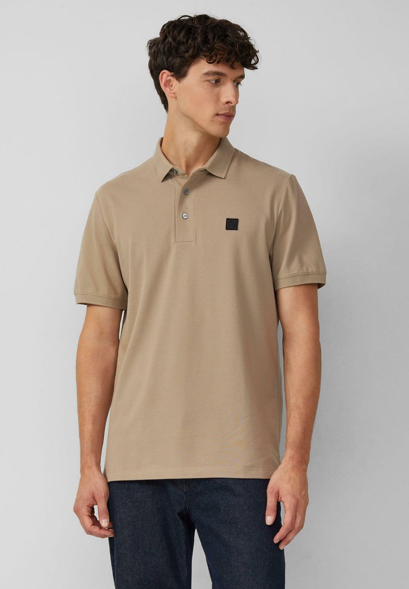 Beige Poloshirt mit klassischem Kragen, drei Knöpfen und schwarzem Logo-Patch. Kurze Ärmel und ein glattes, strukturiertes Baumwollgewebe.
