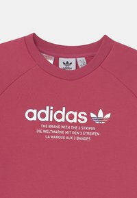 Sudadera rosa con cuello redondo y mangas raglán. Logo y texto en blanco de Adidas en la parte frontal. Material de mezcla de algodón con una textura suave.