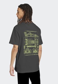 Άνδρας με κοτσίδες, που φοράει μια μαύρη μπλούζα Adidas με κίτρινες τεχνικές σχεδιάσεις ενός αγωνιστικού αυτοκινήτου και την επιγραφή "Σχεδιασμένο για ταχύτητα" στην πλάτη.