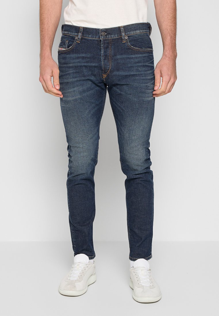 Mann in slim-fit blauen Jeans mit dezentem Verblassen, kombiniert mit weißen Sneakers und einem weißen T-Shirt, vor einem hellen einfarbigen Hintergrund.