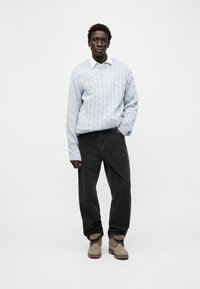  Νεαρός άνδρας όρθιος με το χέρι στην τσέπη, φορώντας ανοιχτό μπλε πουλόβερ με πλέξη καλώδιο, λευκό πουκάμισο με γιακά, μαύρα τζιν και μπεζ μποτάκια, σε απλό φόντο.