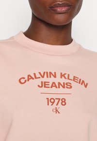 Sudadera de color rosa claro hecha de un tejido suave, con un cuello redondo y el texto "CALVIN KLEIN JEANS 1978" en color óxido.