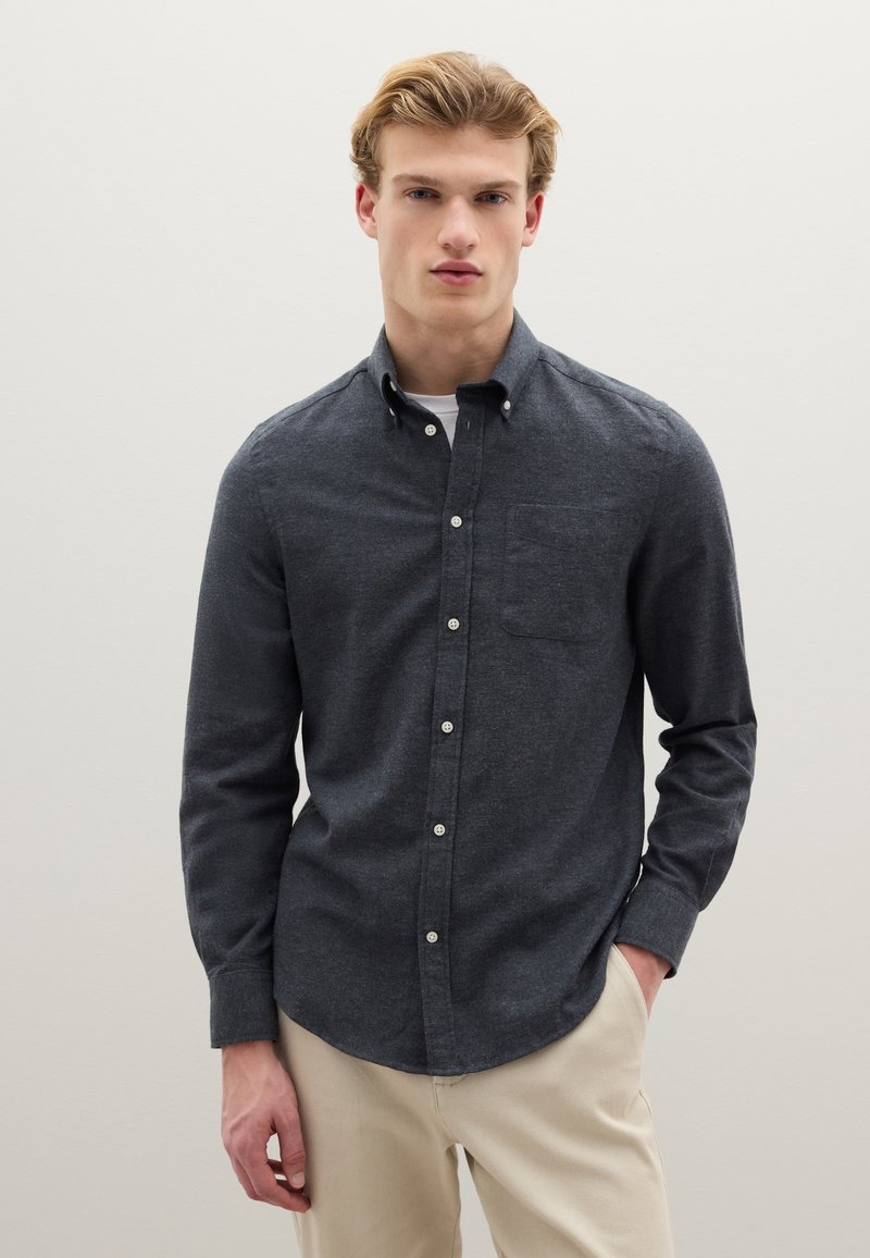 Camisa azul oscuro de manga larga hecha de tela texturizada con un bolsillo en el pecho, botones blancos y dobladillo curvado, combinada con pantalones de color claro.