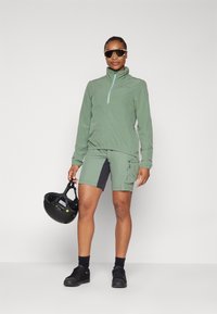 Šviesiai žalia zip-up striukė su elastingomis manžetėmis, derinama su atitinkamomis šortais, kurių šoninės dalys tamsesnės. Užsidėjęs akinius nuo saulės ir laiko juodą šalmą.