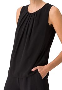 Femme portant un chemisier noir sans manches avec des plis au niveau de l'encolure, main dans la poche, recadrée aux hanches.