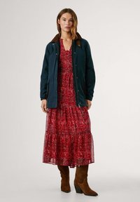 Marineblauwe jas met een bruine kraag, gedragen over een rood gepatterne maxi-jurk. Bruine haklaarzen maken de outfit compleet.