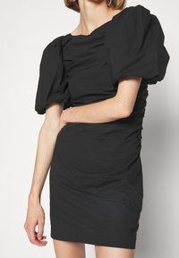 Svart klänning med puffärmar och rynkdetaljer. Kroppsnära passform, slät textur och fyrkantig halsringning. Kort nederdel, ingen synlig hårdvara.