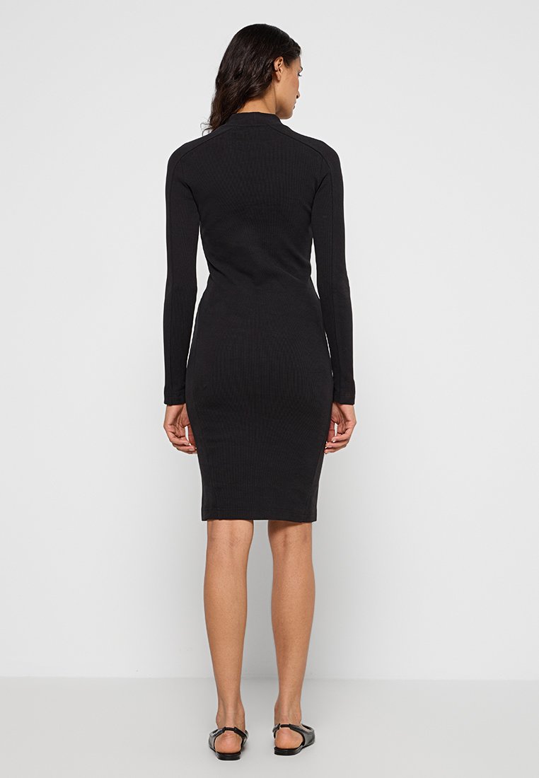 Femme debout de dos, portant une robe noire ajustée à manches longues et des sandales plates noires, sur fond blanc uni.