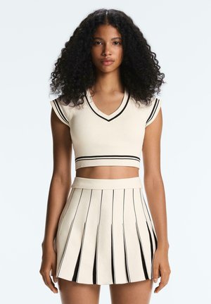 Kobieta o kręconych włosach, ubrana w kremowy krótki top i plisowaną spódnicę z czarnymi pionowymi paskami, stojąca na tle jednolitego tła.