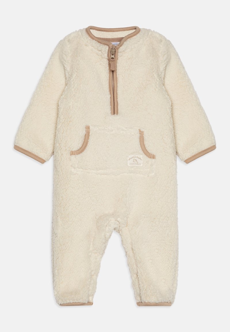 Combinaison pour bébé en polaire couleur crème avec manches longues, fermeture éclair à l'avant, bordure marron et une poche frontale avec une petite étiquette.