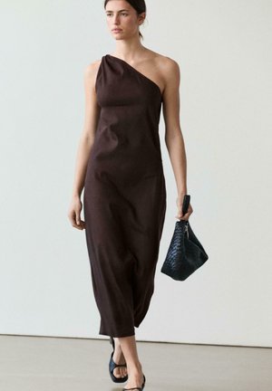 Frau geht, trägt ein ein-schulteriges dunkelbraunes Kleid, schwarze offene Absatzschuhe und trägt eine schwarze strukturierte Handtasche.
