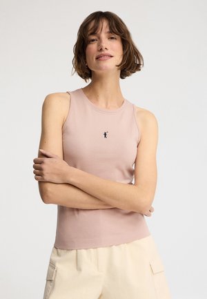 Femme aux cheveux courts et bruns, portant un haut sans manches rose clair avec un petit logo foncé et un pantalon clair, bras croisés.