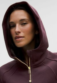 Bordo zip-up kapuca s teksturirano tkanino, z zlatim zadrgo, visokim ovratnikom in kapuco na vrvico. Posnetek oblačila od blizu.