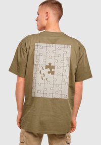 Oliven grøn bomulds t-shirt med et puslespilsbrik grafik på ryggen, der viser en manglende brik og et givende printdesign.