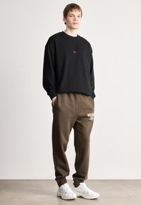 Μαύρος φούτερ με μικρό κόκκινο λογότυπο, καφέ στενός ιδρώτας με λευκή γραφή, και λευκά αθλητικά παπούτσια σε ουδέτερο φόντο.