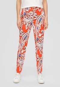 Pantalons rouges à motifs avec des designs en paisley blancs, bleus et verts, coupe slim, taille élastique et longueur jusqu'à la cheville, associés à des baskets blanches.