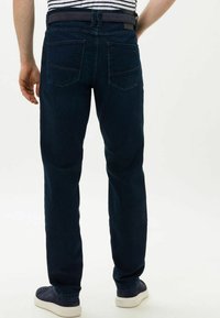 Donkerblauwe jeans van denim, met een recht gesneden ontwerp, twee achterzakken en een zwarte riem met een lus. Draagt met lichtgekleurde sneakers.