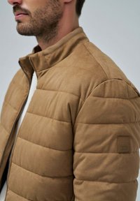 Chaqueta acolchada en tono marrón con tejido suave y texturizado, cuello alto y cremallera. Presenta costuras horizontales y un pequeño parche con el logotipo en la manga.