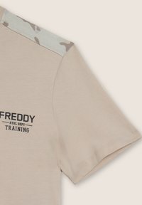 T-shirt beige a maniche corte, con un accento in camouflage sulla spalla e testo stampato in nero che recita "FREDDY ATHL DEPT TRAINING."