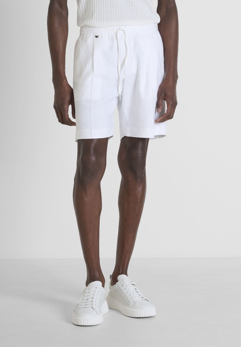 Shorts en lin blanc avec taille élastique et cordon de serrage. Texture lisse et plis sur les côtés. Associés à des baskets blanches.