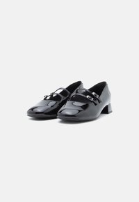 Par de sapatos Mary Jane pretos em couro envernizado com saltos baixos e tiras duplas com fivela, num fundo branco.