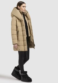 Beige quiltet pufferjakke med høj krave, lynlås foran og sorte detaljer. Båret over sorte tætsiddende leggings og kraftige sorte støvler.